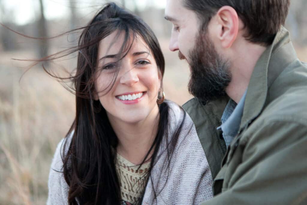 Candid Colorado engagement photo. Close-up photo of a woman smiling at the camera as the wind blows her hair, sitting with her partner outdoors in a grassy field.