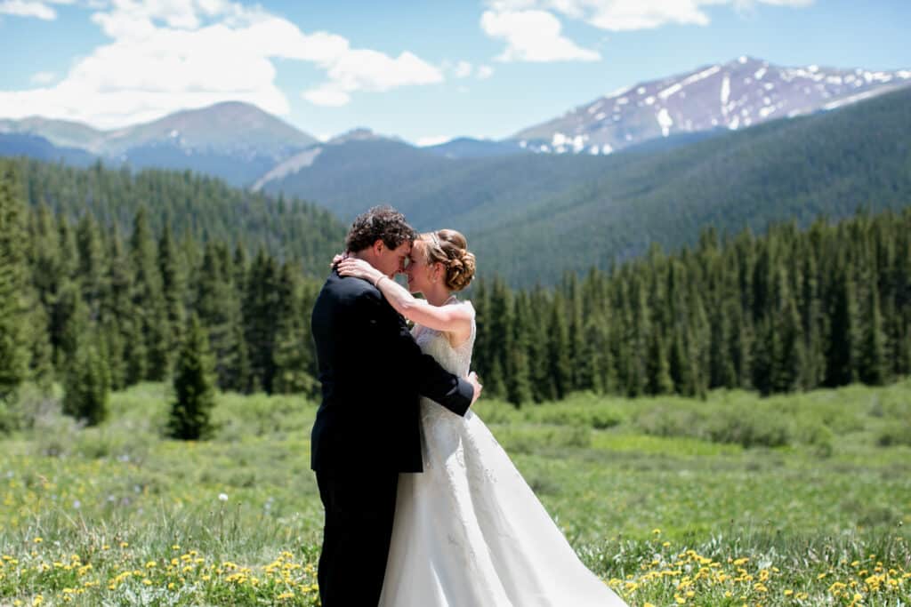 Breckenridge Colorado mountain wedding portrait. Bride and groom embracing during their wedding in an alpine meadow with evergreen trees and snow-capped mountains in the background in Breckenridge, Colorado.
