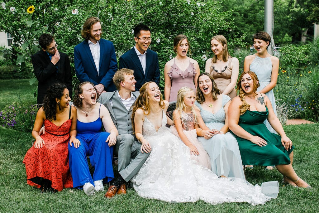 Candid Colorado wedding party photo. Bride and groom sitting on the grass surrounded by their wedding party, all laughing together during an outdoor wedding celebration.