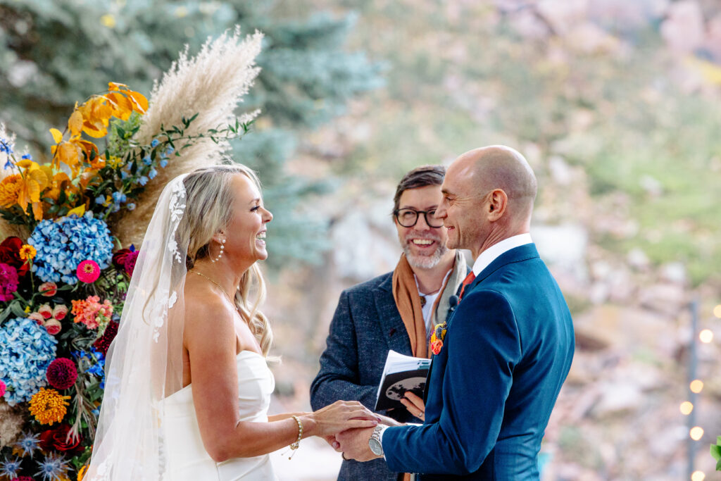 River Bend Lyons Colorado wedding ceremony. Bride and groom smiling at each other during their wedding ceremony at River Bend in Lyons, Colorado, with their officiant behind them and colorful florals in the background.