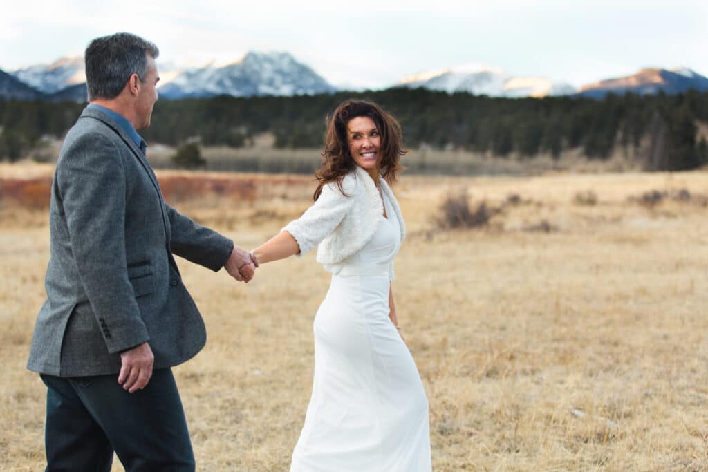 Rocky Mountain National Park elopement photo. Bride smiling over her shoulder while holding hands with her partner during an elopement in a mountain meadow near Rocky Mountain National Park, with snow-covered peaks in the background.