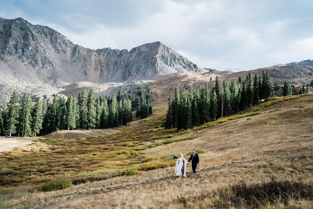 Colorado mountain wedding at Arapahoe Basin. Bride and groom walking across a wide alpine meadow at Arapahoe Basin with dramatic mountain peaks and evergreen trees in the background.